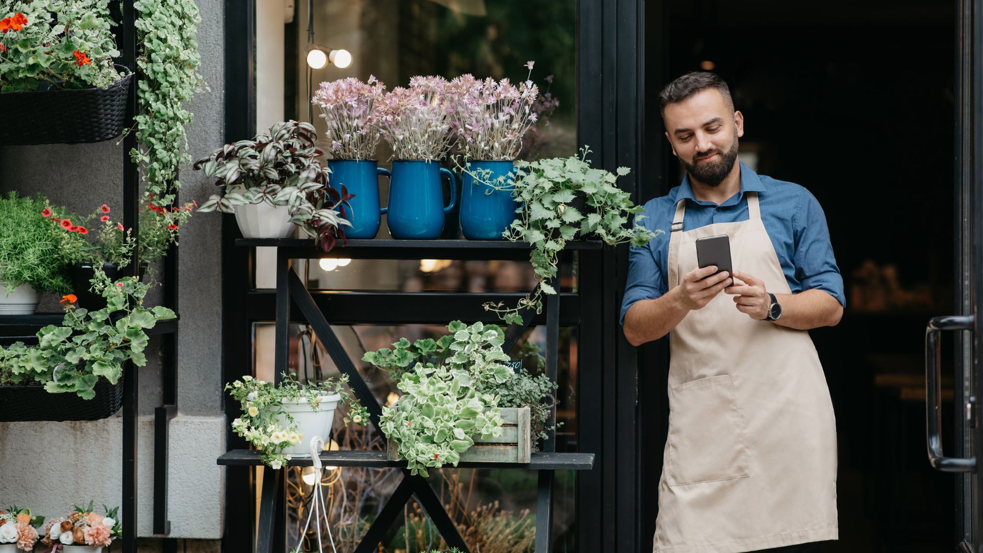 A man in an apron is using his phone outside of a flower shop.